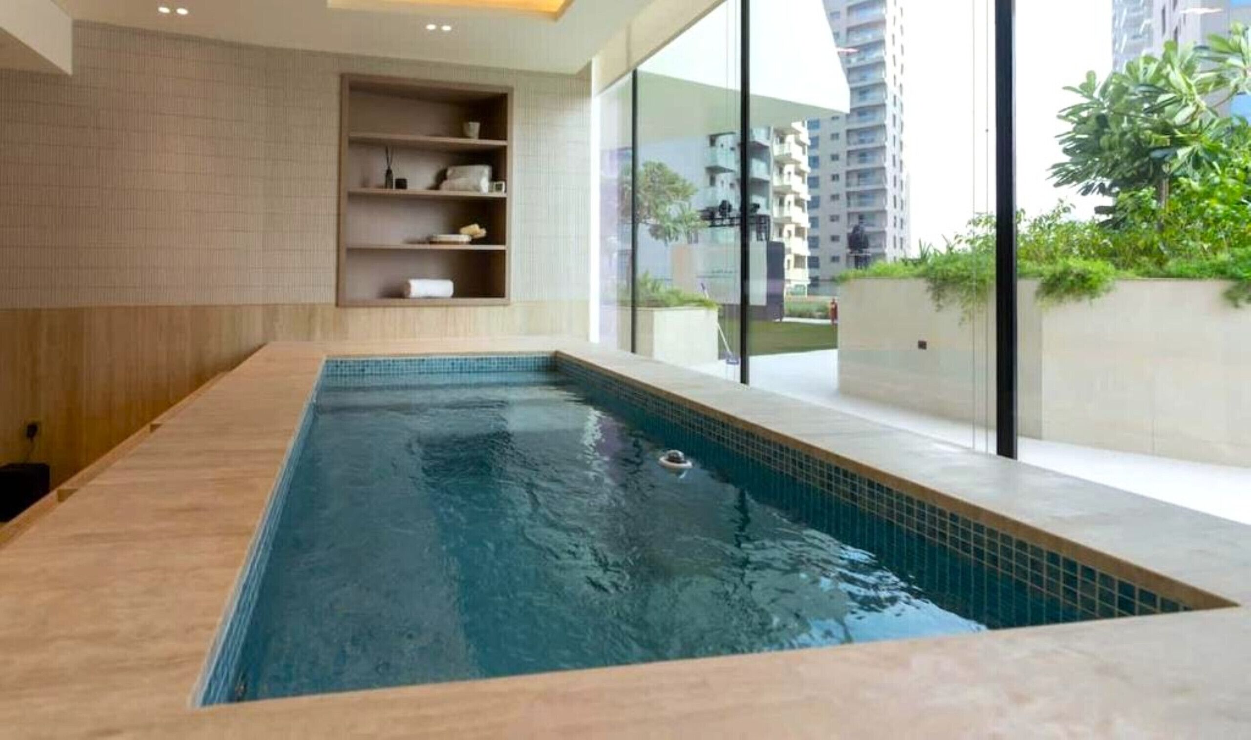 Una piscina interior moderna con paredes de vidrio transparente, suelo de madera y un estante empotrado con toallas.