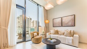 Een moderne woonkamer met een bank, fauteuil, salontafel, grote ramen met gordijnen, en twee abstracte schilderijen aan de muur.