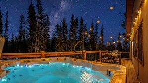 Een hottub met een houten terras, omringd door hoge bomen en een sterrenhemel.