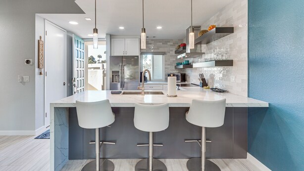 Een moderne keuken met een marmeren aanrecht, drie witte barkrukken, roestvrijstalen apparatuur en hanglampen.