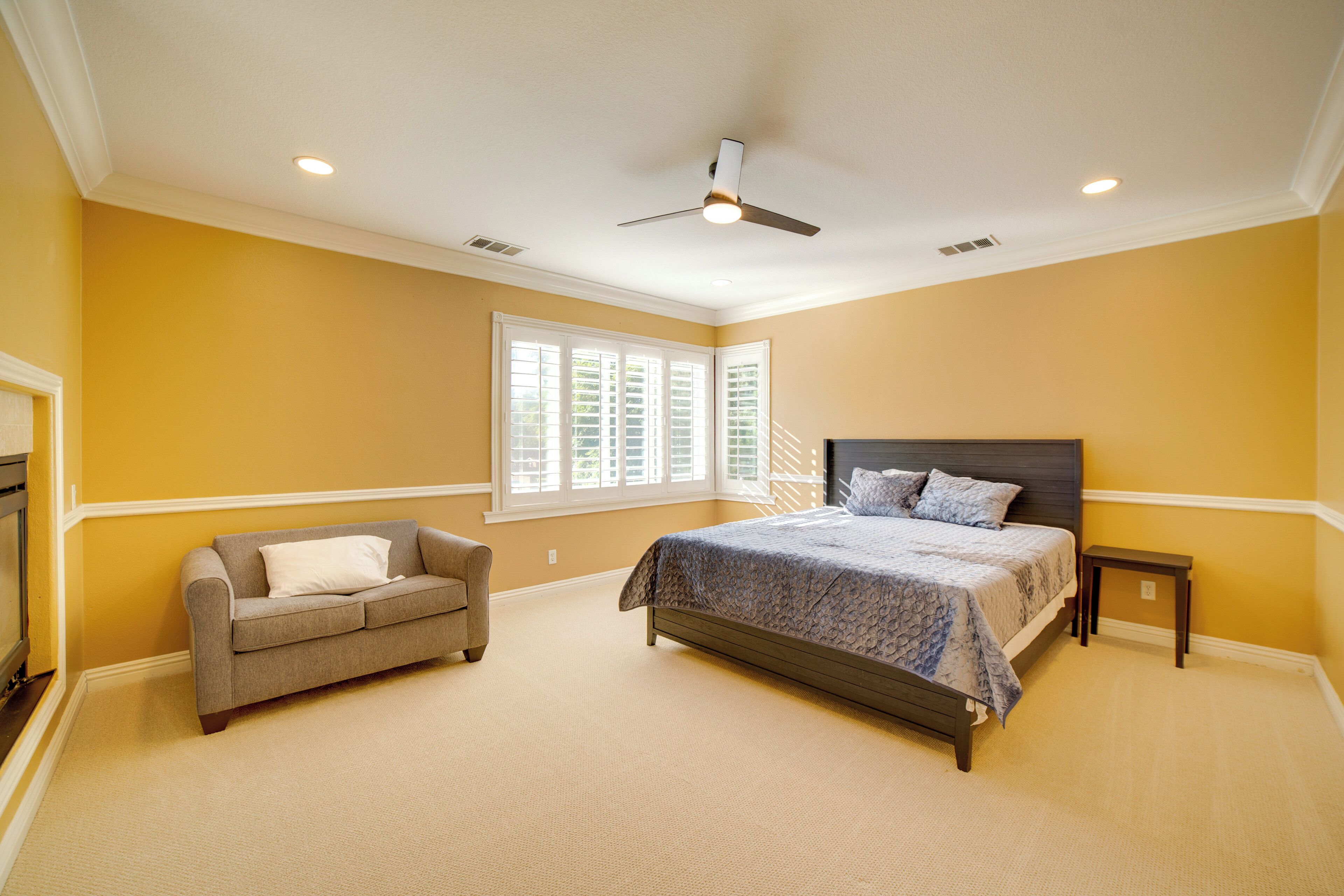 A bedroom with a bed, a sofa, a ceiling fan, and a window with shutters.