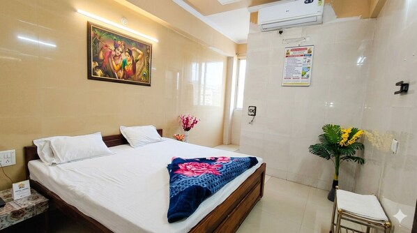 A hotel room with a bed, a nightstand, a wall-mounted air conditioner, a painting, a poster, and a potted plant. - SHRI HARIVANSH SEWA KUNJ - Near Banke Bihari Temple Vrindavan (Vrindavan)
