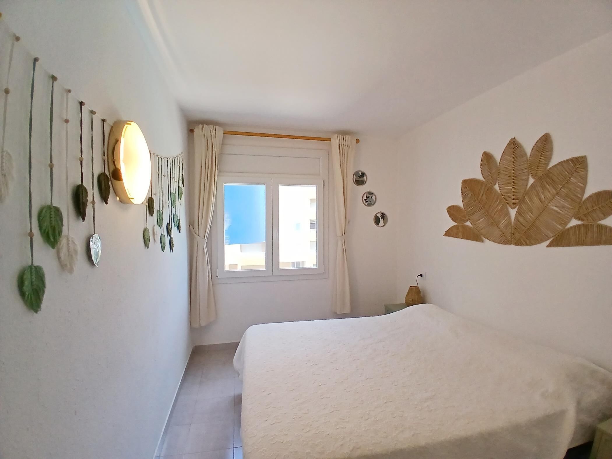 A bedroom with a bed, a window with curtains, a wall decoration, and a wall-mounted lamp.