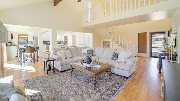 A spacious living room with a large sectional sofa, a glass coffee table, and a wooden floor. A staircase leads to an upper level with a balcony. - Groups Welcome, Central Location! Tulsa Gem + Yard (Tulsa)