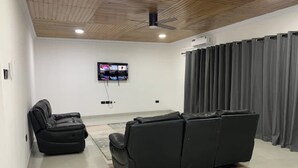 A modern living room with a sectional sofa, a flat-screen TV mounted on the wall, and a ceiling fan. - Tree fort property 2 in North Legon (Accra)