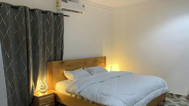 A single bed with a wooden headboard, a nightstand with a lamp, a patterned curtain, and a tiled floor. - Tree fort property 2 in North Legon (Accra)
