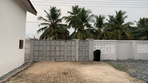 A paved driveway leading to a gated entrance with a white wall and palm trees in the background. - Tree fort property 2 in North Legon (Accra)