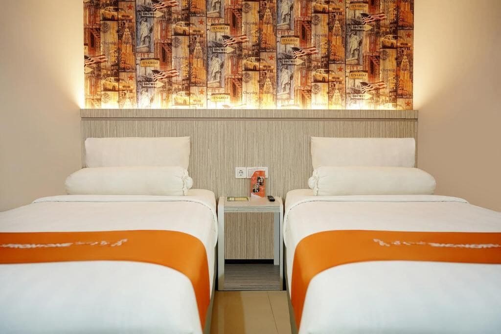 Habitación de hotel con dos camas, cada una con ropa blanca y detalles naranja, una mesita de noche pequeña con lámpara y una pared con papel tapiz de estilo vintage.