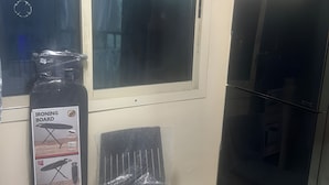 A room with a large window, a black ironing board, a small chair, and a black cabinet. - OST Diera Market Homes (Dubai)