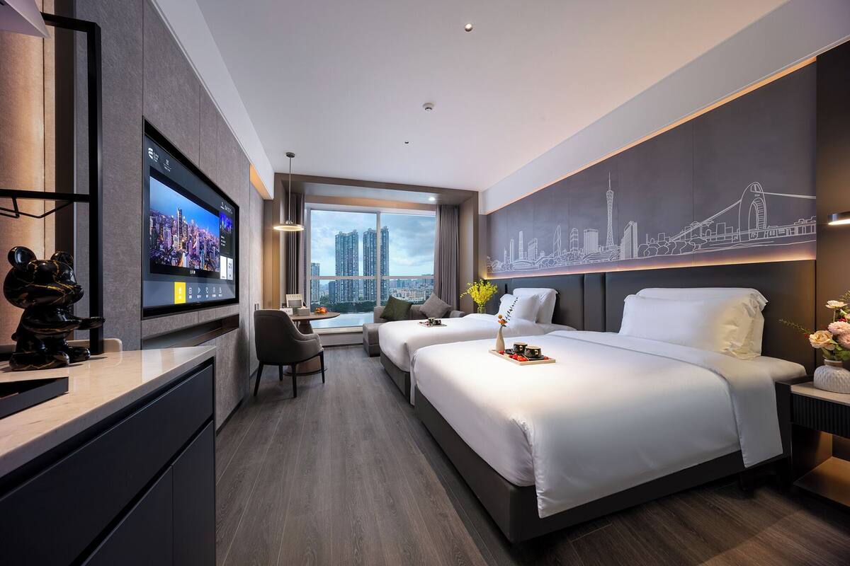 Habitación de hotel moderna con un televisor grande, dos camas, un escritorio y vistas a la ciudad.