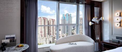 A hotel room with a large window, a curved sofa, a bedside table with a fruit bowl, and a lamp.