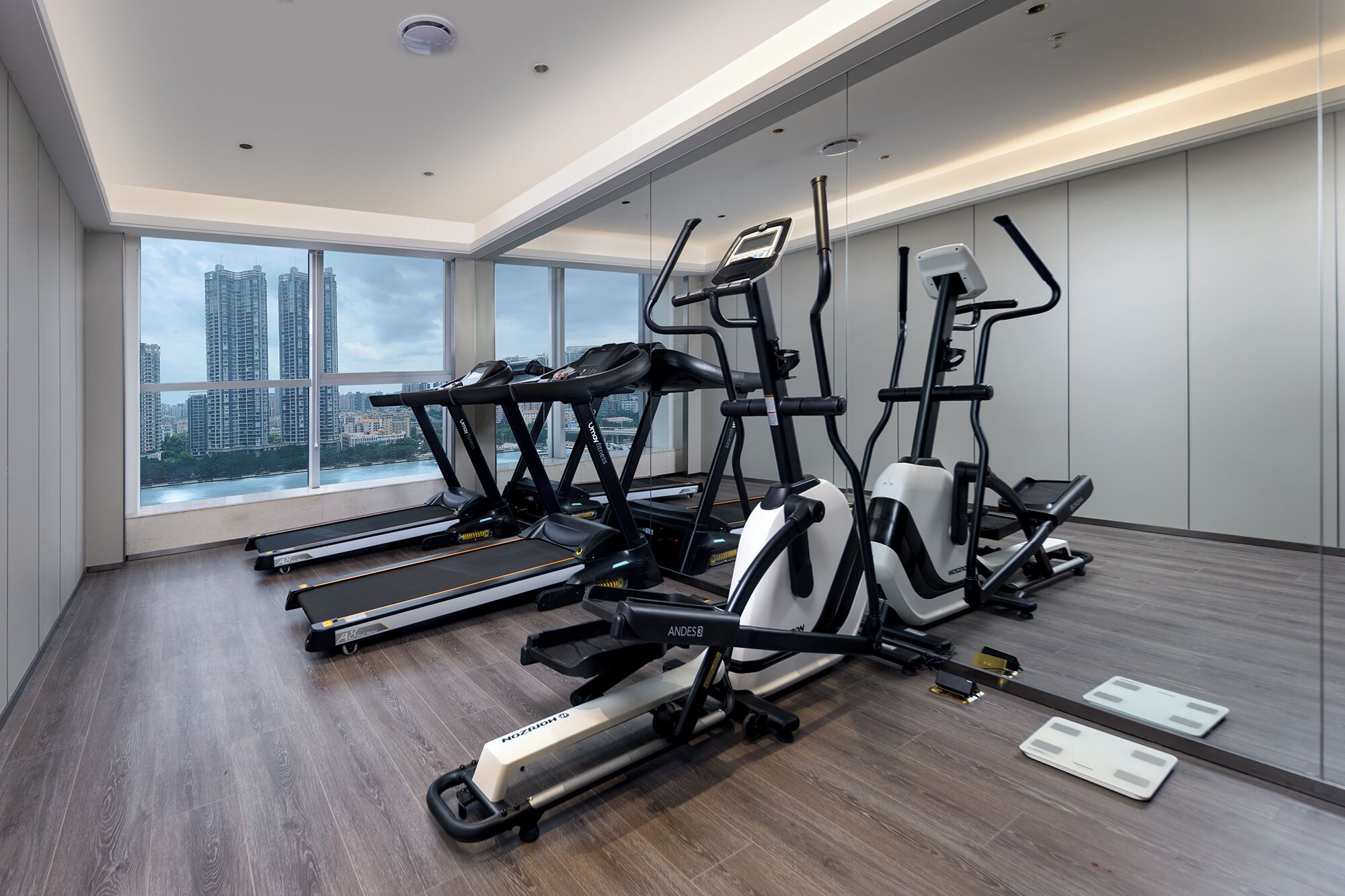 A modern gym with multiple cardio machines, large windows offering a city view, and a wooden floor.