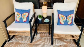 Two wicker chairs with white cushions and blue butterfly pillows, a small table with a lamp, a cup, and a plant, and a patterned rug. - Cozy King Size 1-bedroom condo in adorable La Quinta with AC (La Quinta)