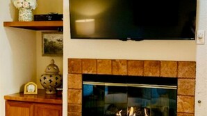 A living room with a fireplace, a television mounted above it, and a wooden cabinet to the left. - Cozy King Size 1-bedroom condo in adorable La Quinta with AC (La Quinta)