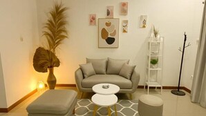 A living room with a sofa, two ottomans, a coffee table, a floor lamp, a bookshelf, and a large plant. - Shaionara Villa (Parongpong)