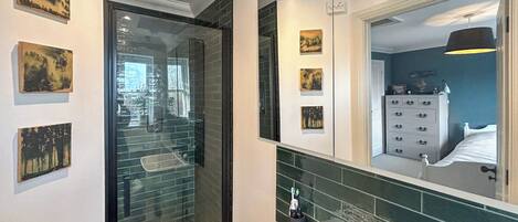 A bathroom with a glass shower enclosure, a white sink, and a mirror reflecting a bedroom with a dresser and a bed.