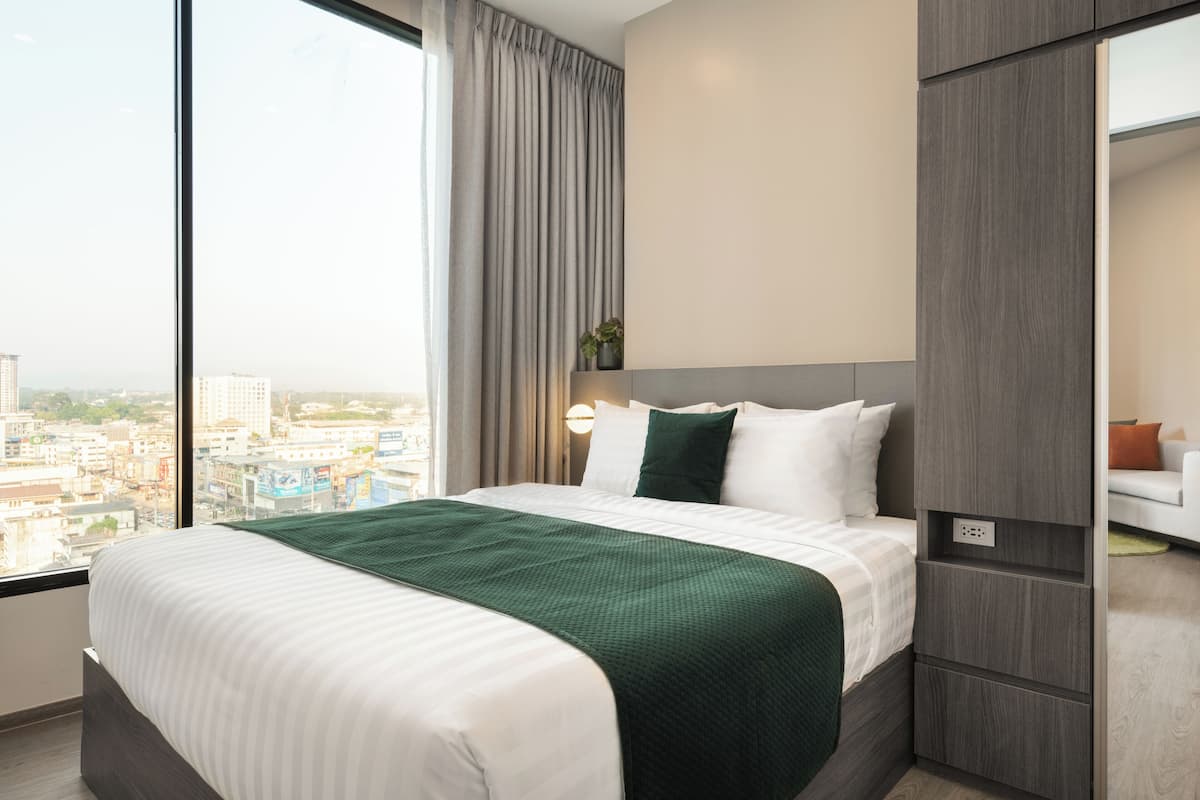 A hotel room with a large window, a bed with a green blanket, a nightstand, and a wardrobe.