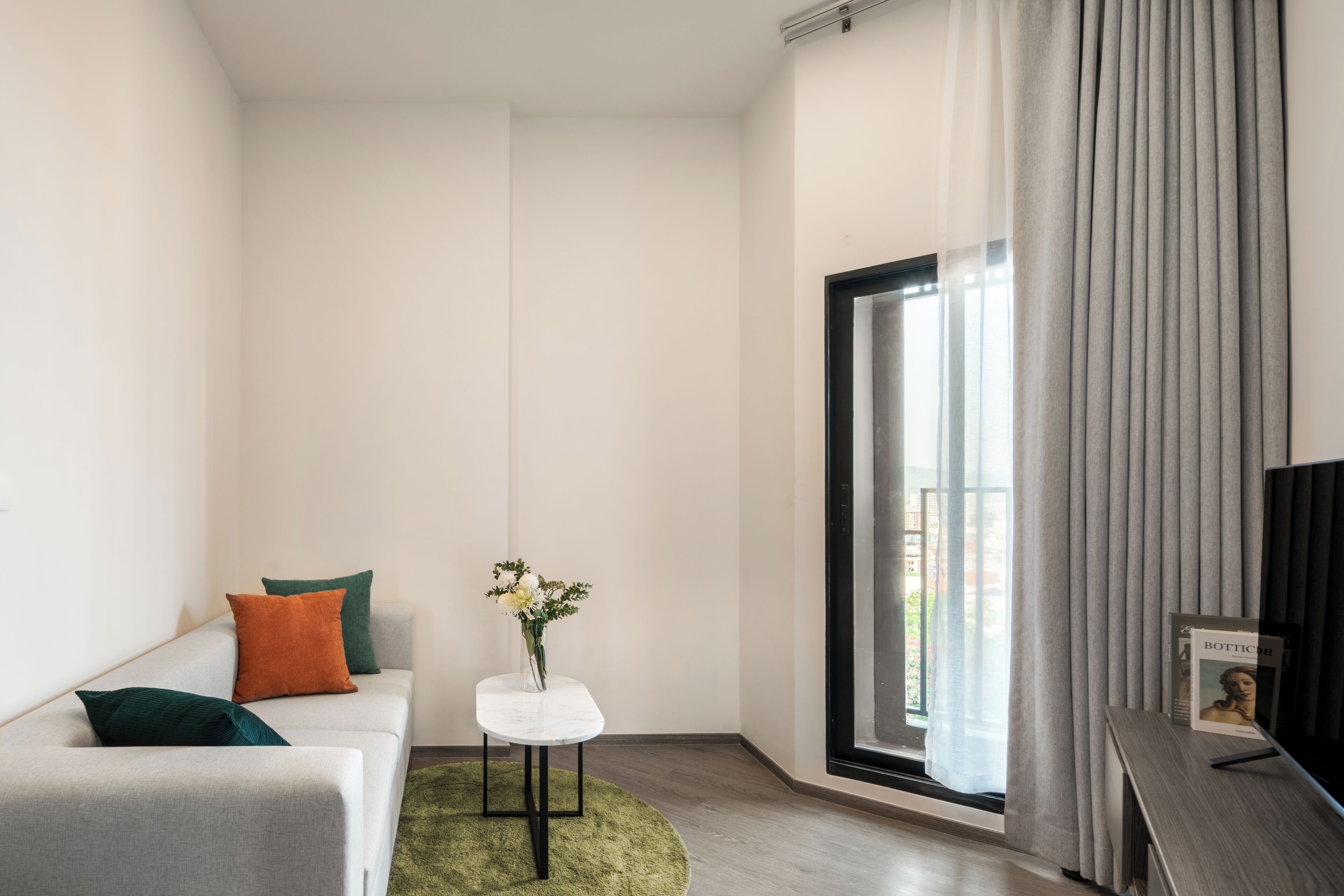 A modern living room with a sofa, a small table with a vase, and a large window with curtains.