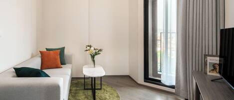 A modern living room with a sofa, a small table with a vase, and a large window with curtains.