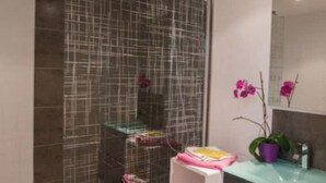 Ein Badezimmer mit Duschabtrennung aus Glas, einem weißen Waschbecken mit einer violetten Pflanze und einem Spiegel darüber.