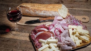 Ein Wurst- und Käsebrett mit verschiedenen Fleischsorten, Käsesorten und Gewürzgurken, dazu ein Brotlaib und ein Glas Rotwein auf einem Holztisch.