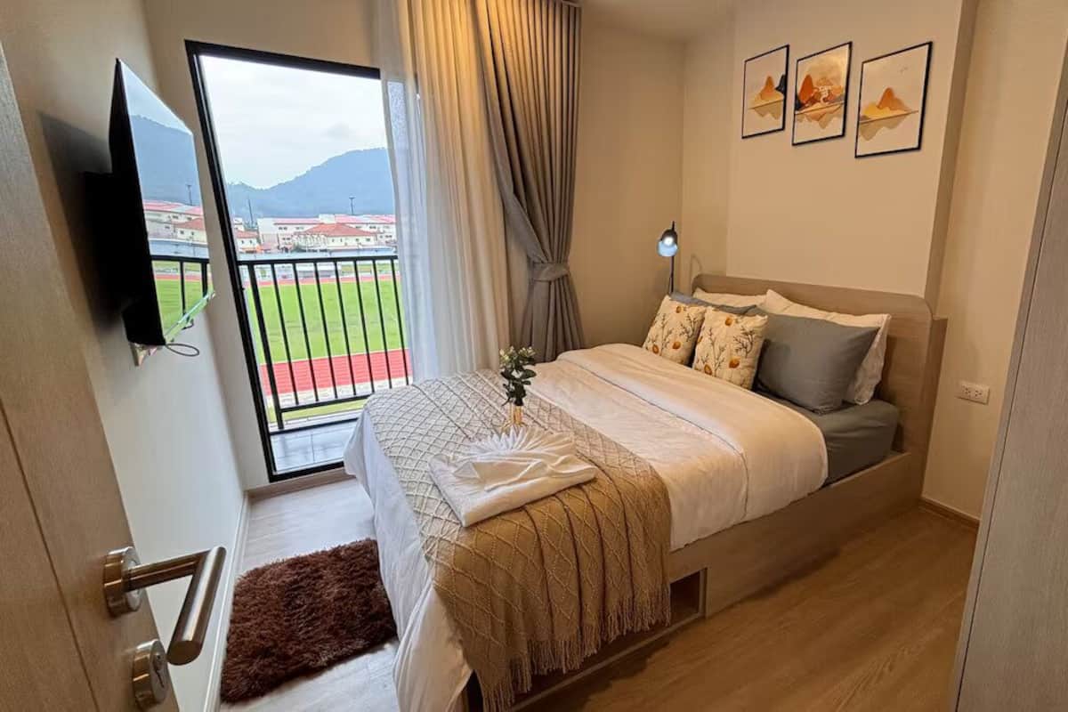 Ein Schlafzimmer mit Bett, Fernseher, Fenster mit Blick auf ein Sportfeld und einem Balkon.