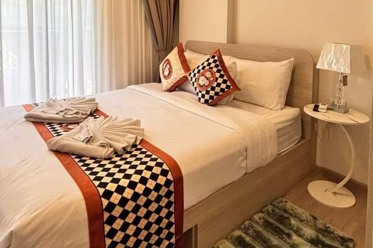 Ein Hotelzimmer mit Bett, Nachttisch, Lampe, einem Fenster mit Vorhängen und einem Teppich.