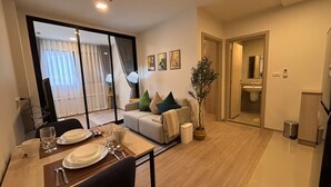 A modern living room with a dining area, a sofa with cushions, a potted plant, and a sliding glass door leading to a balcony. - 2 BR Dcondo Reef Phuket By Ploy (Kathu)