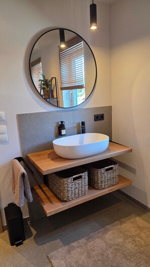 Une salle de bain avec un miroir rond, un lavabo blanc, des étagères en bois, des paniers en osier et un porte-serviettes.