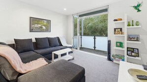 Ein modernes Wohnzimmer mit einem grauen Sofa, einem weißen Couchtisch und einem großen Fenster mit Blick auf Grünflächen.