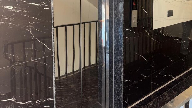 A marble staircase with a glass elevator door, a light fixture on the wall, and a metal railing. - Hotel Rajshree (Hisar)