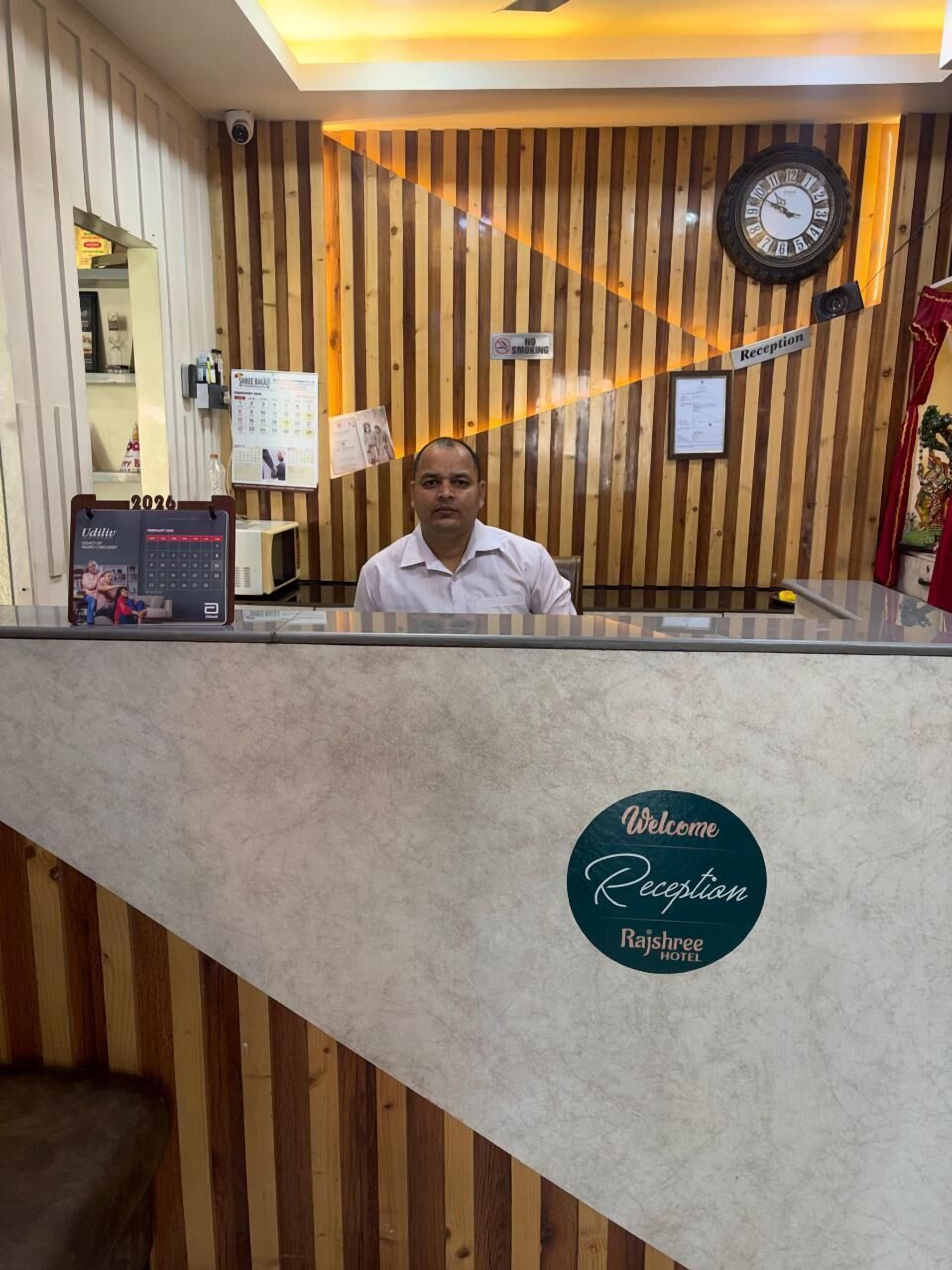 Mostrador de recepción con un reloj, un letrero de 'No Fumar', un calendario y un cartel de 'Welcome Rajshree Hotel'.