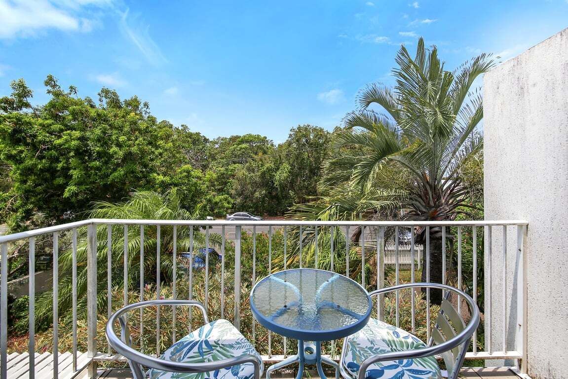 A balcony with a glass table and two chairs, surrounded by lush greenery and a palm tree.
