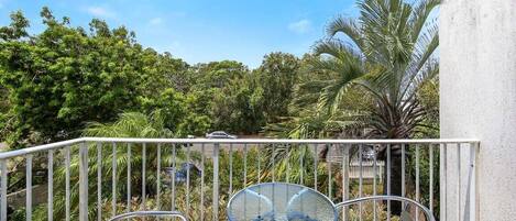 A balcony with a glass table and two chairs, surrounded by lush greenery and a palm tree.