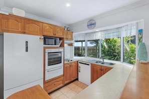 Una cocina con armarios de madera, un refrigerador blanco, un microondas, un horno, un lavavajillas y una ventana con vistas a la vegetación.