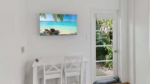 Een kamer met een witte muur, een televisie boven een witte tafel met twee stoelen, en een deur die leidt naar een tuin met tropische planten.