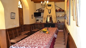 Un coin repas avec une longue table dressée pour le repas, des bancs en bois le long des murs, une cheminée et, au-dessus, un crâne de cerf accroché au mur.