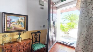Ein Zimmer mit einem Holzkasten, einem Sessel, einem Gemälde und einer Tür, die auf einen Balkon mit Pflanzen führt.