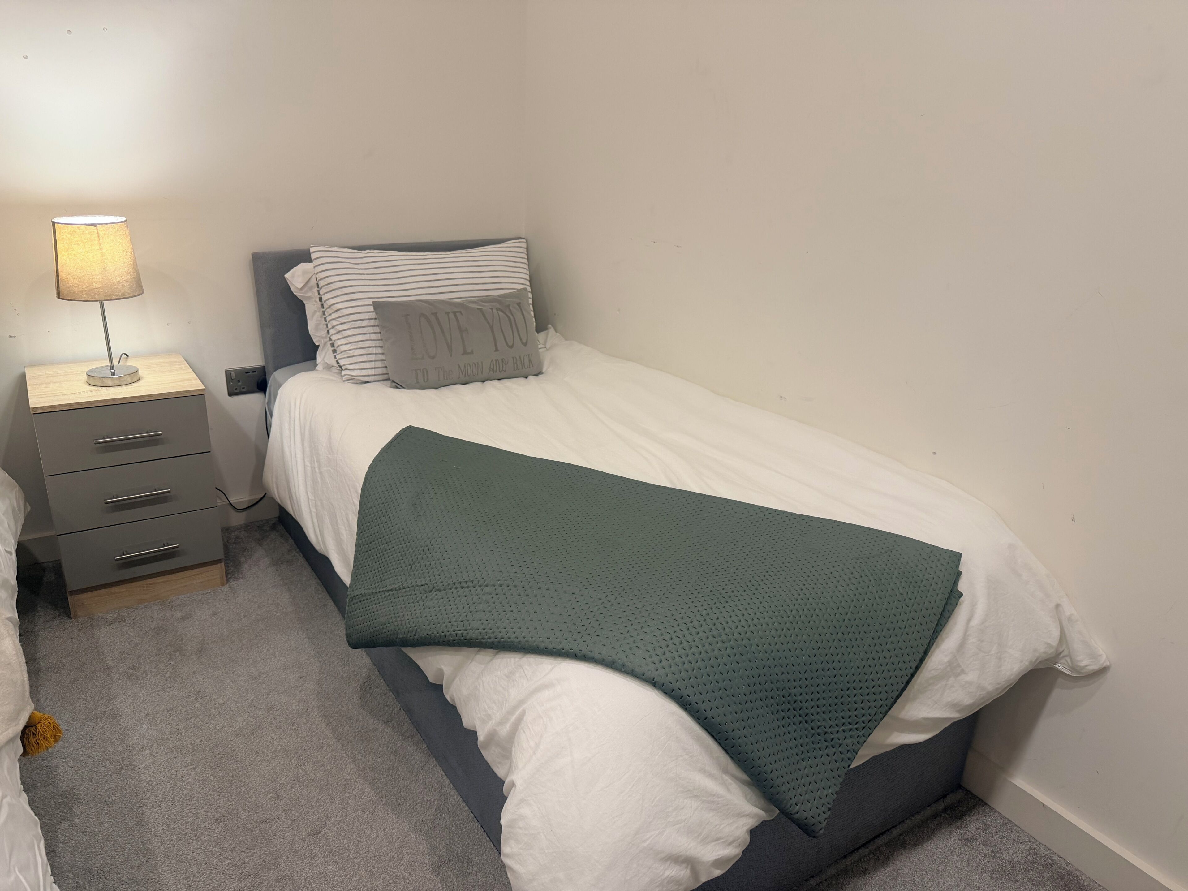 A bedroom with a single bed, a bedside table with a lamp, a drawer unit, and a green blanket.