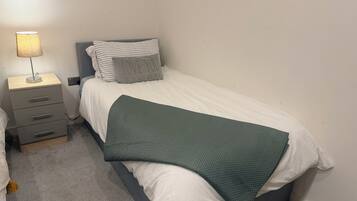 A bedroom with a single bed, a bedside table with a lamp, a drawer unit, and a green blanket.
