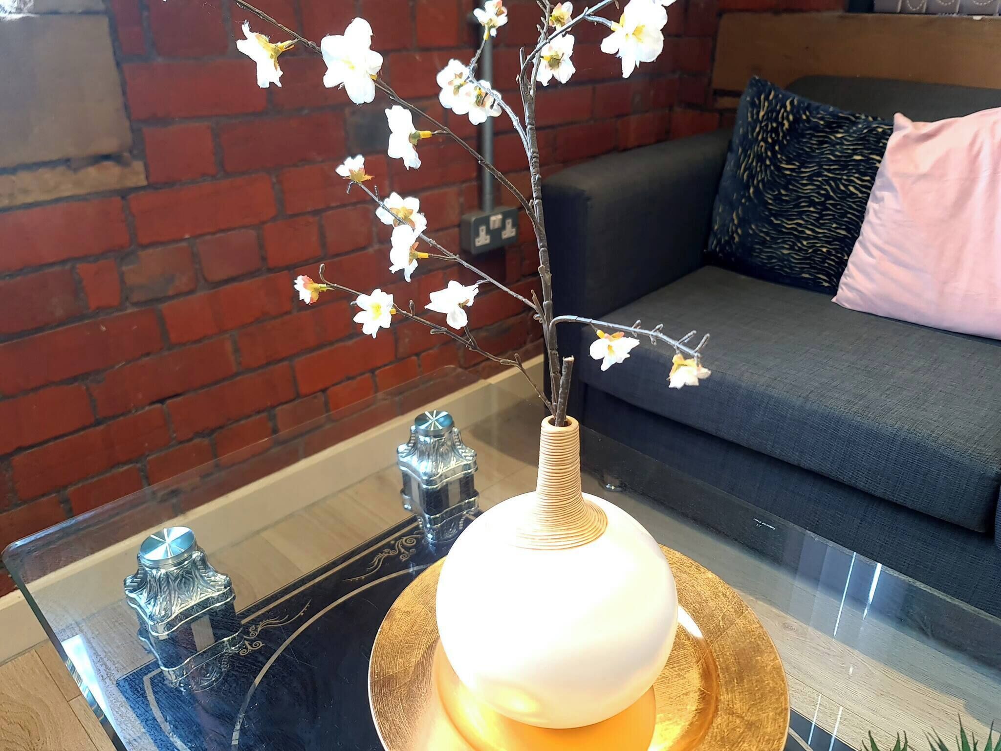 Una mesa de vidrio con un florero de flores blancas, un objeto metálico decorativo y un cojín en un sofá.