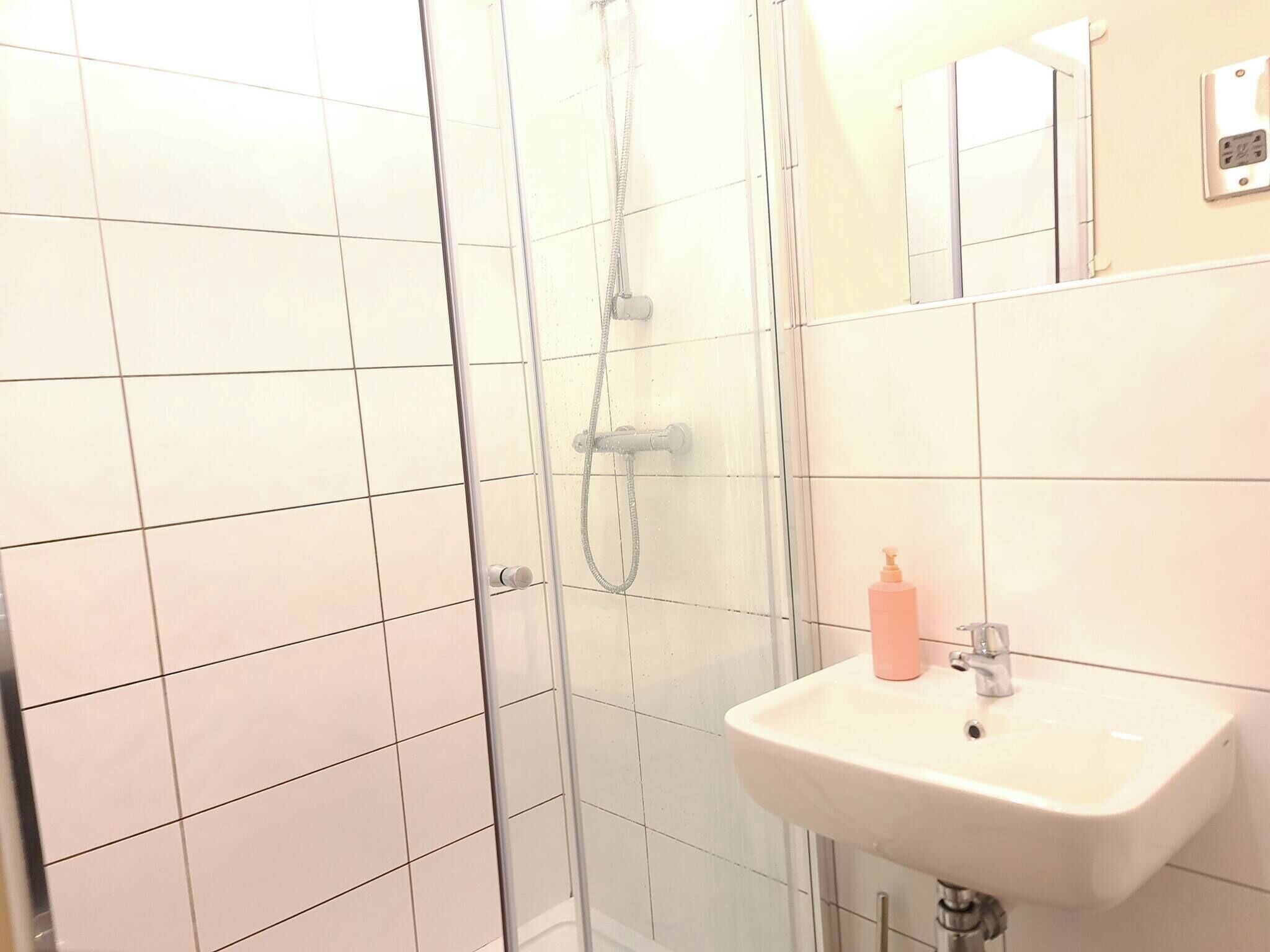 A bathroom with white tiled walls, a glass shower enclosure, a sink with a mirror, and a bottle of soap.