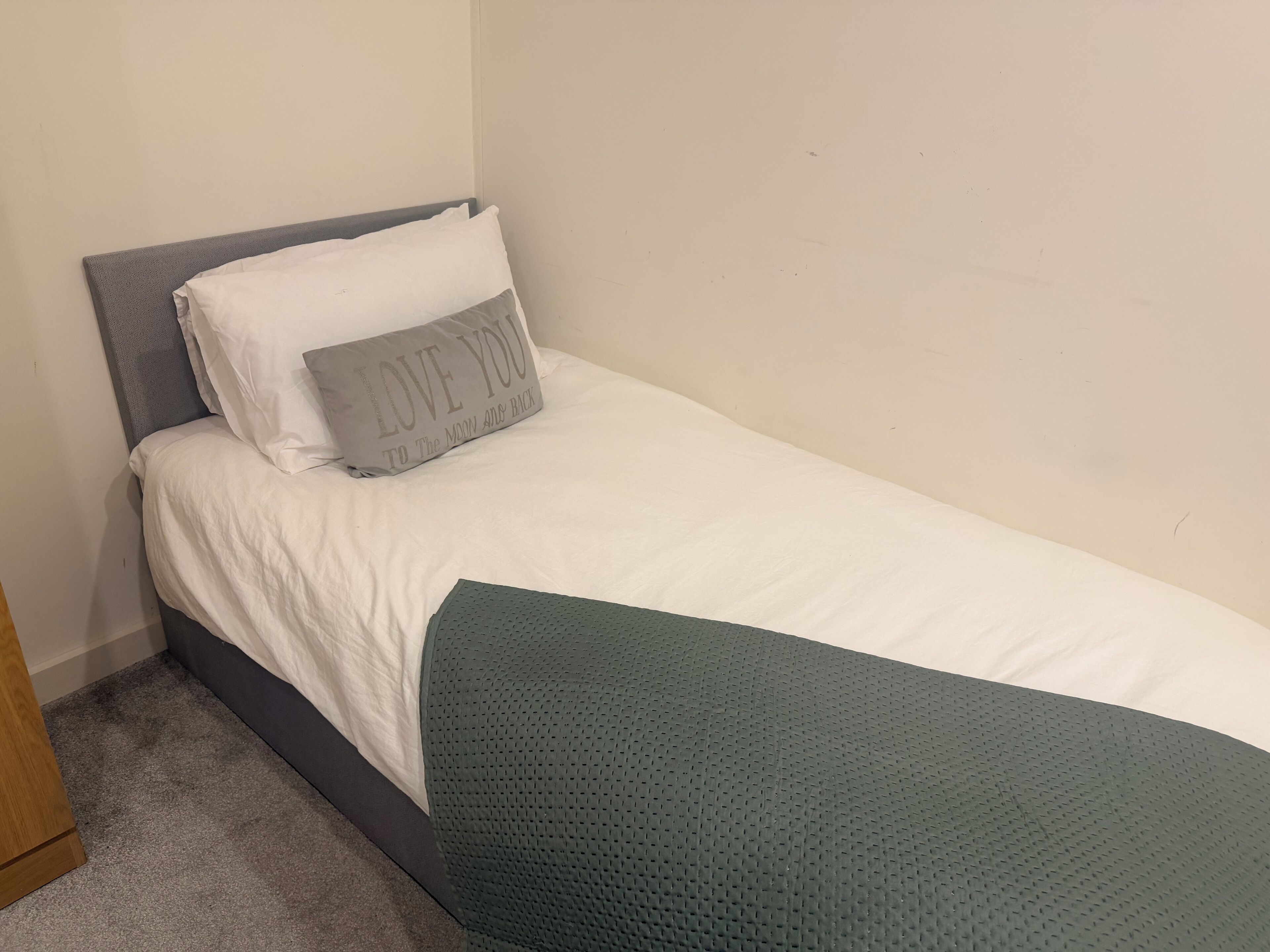 A single bed with a grey headboard, white bedding, and a green blanket.