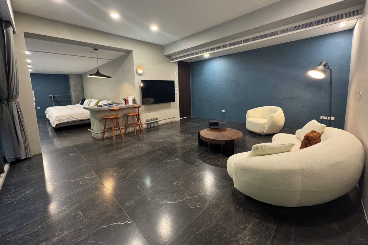 A modern hotel room with a large bed, a seating area with a sofa and chairs, a small table, and a TV mounted on the wall.