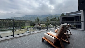 Varanda com duas cadeiras de descanso e uma mesa, com vista para uma paisagem montanhosa e um estacionamento.