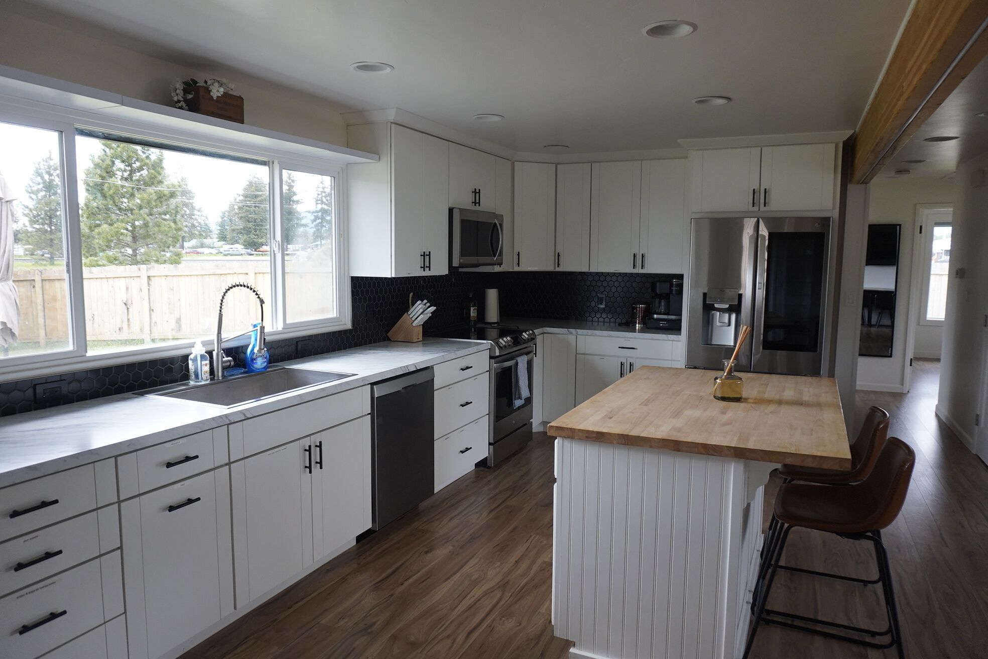 A modern kitchen with white cabinets, a wooden countertop, a stainless steel refrigerator, and a window with a view of trees.