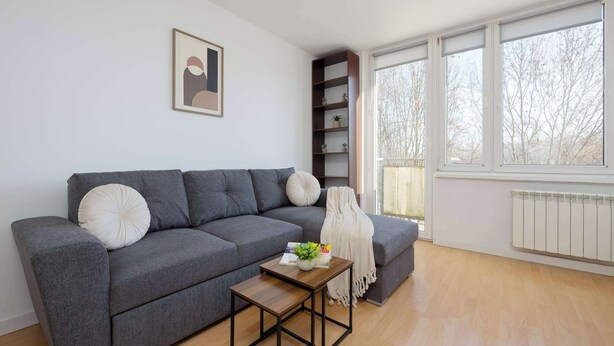 En moderne stue med en grå sofa, et lille træbord, en reol og et vindue med udsigt til træer.