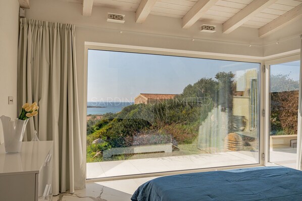 A bedroom with a large sliding glass door leading to a balcony with a view of a hillside and a building.