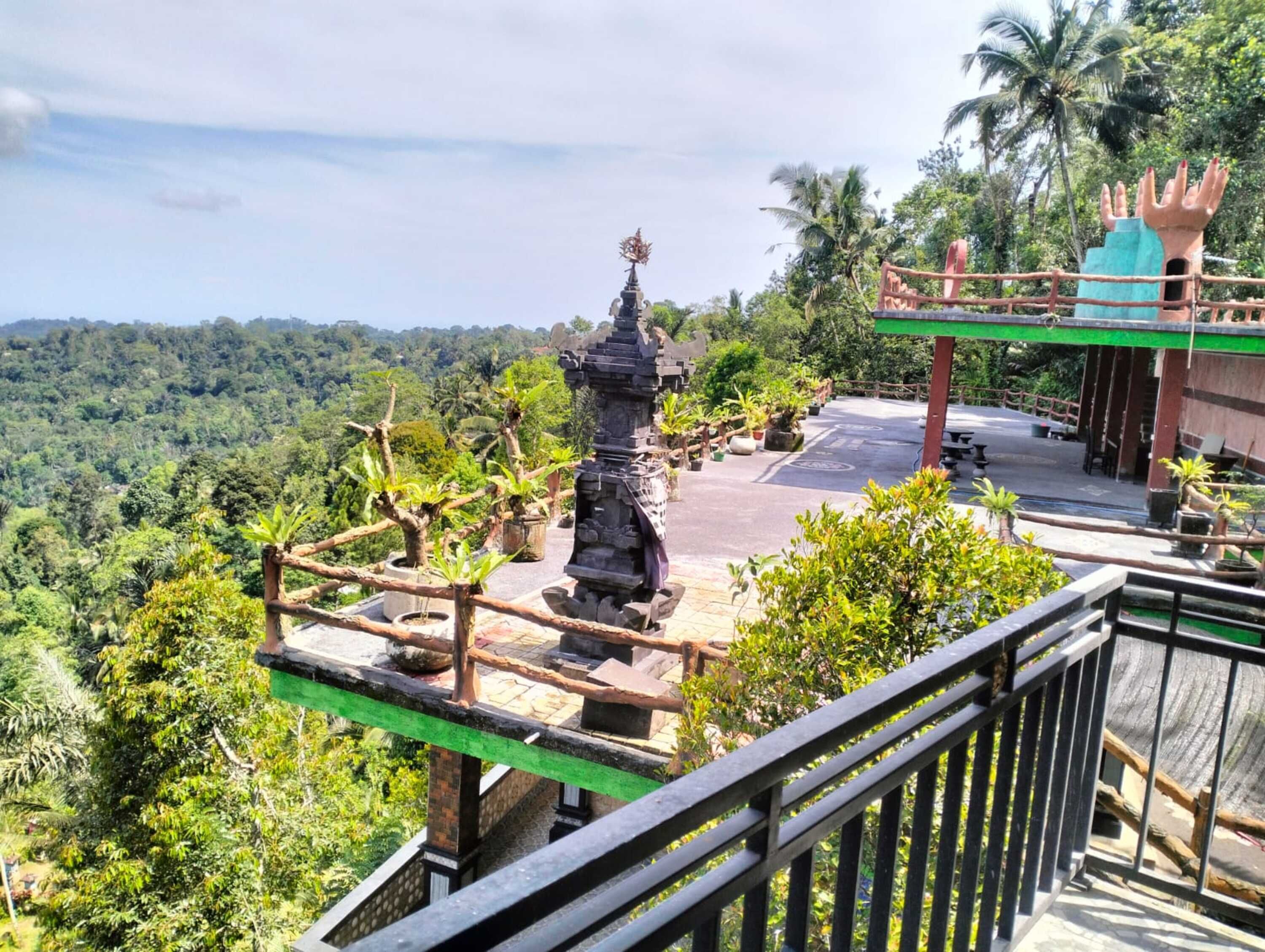 Vista desde un balcón sobre un paisaje exuberante con una estatua de piedra, una cerca de madera y un edificio con techo verde.