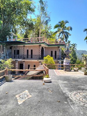 A two-story building with a balcony, surrounded by lush greenery and palm trees.